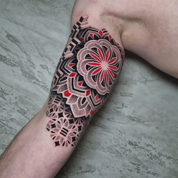 Intricate mandala tattoo in red and black ink on upper arm, showcasing detailed geometric patterns.