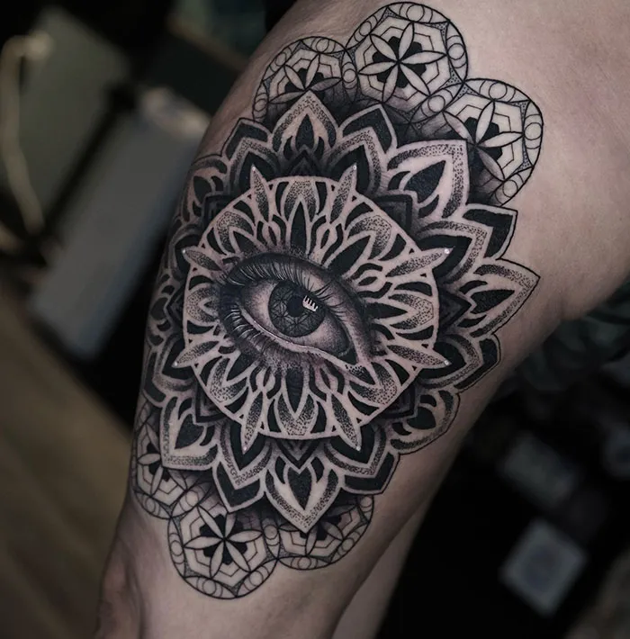 Intricate mandala tattoo featuring a central eye design on the thigh, showcasing detailed patterns and shading.