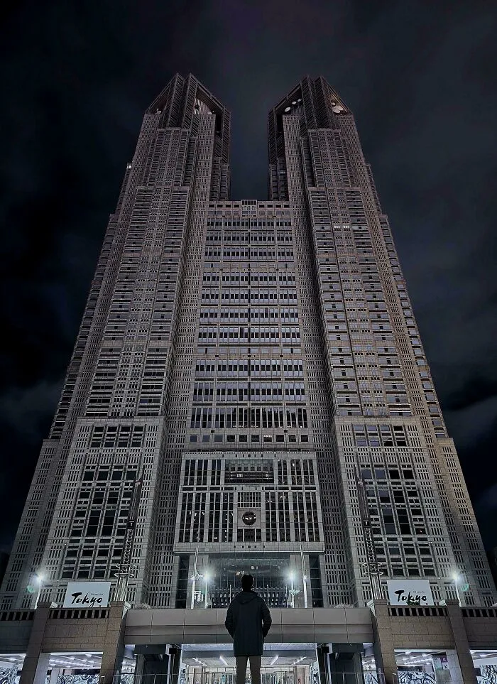 Man standing in front of a towering building with an evil aura, resembling a supervillain headquarters at night in Tokyo.