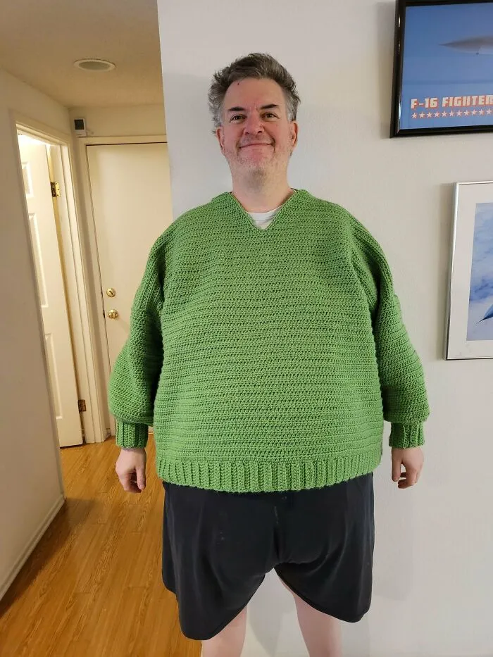 Man wearing a green crocheted sweater, standing indoors, showcasing homemade knitting creativity.