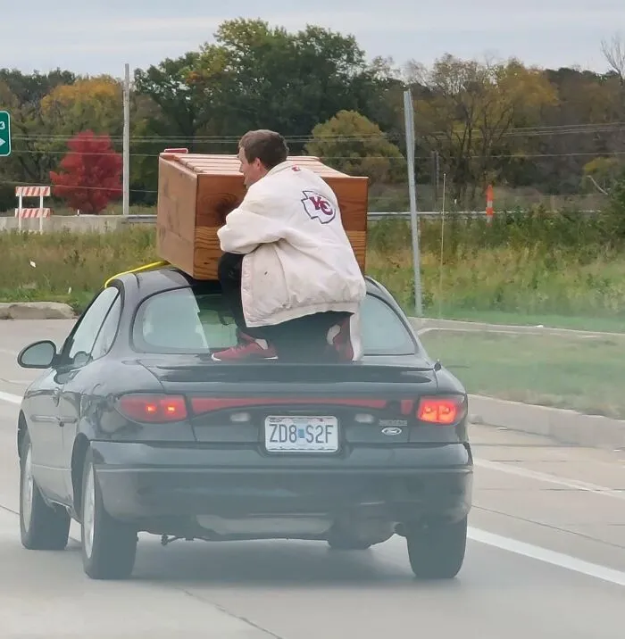 Guy Riding Down The Road Holding A Dresser On Top Of A Car On A 50 Mph Road