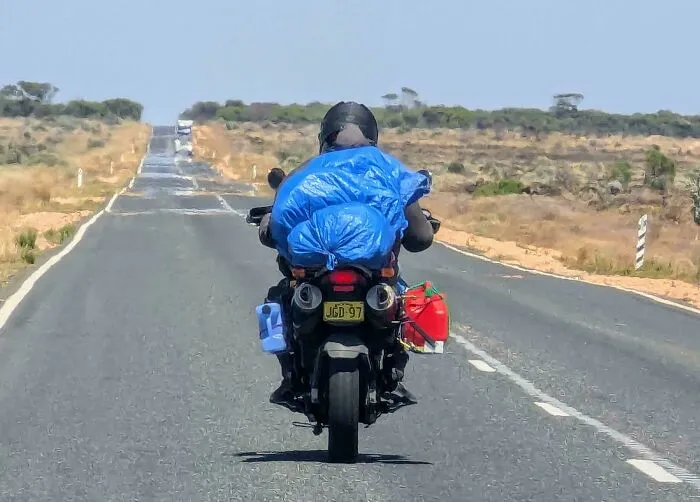 Alguien conduce a través de Nullarbor con su pequeña motocicleta, sus pertenencias en una bolsa de plástico azul y un bidón de combustible de repuesto atado a la motocicleta.