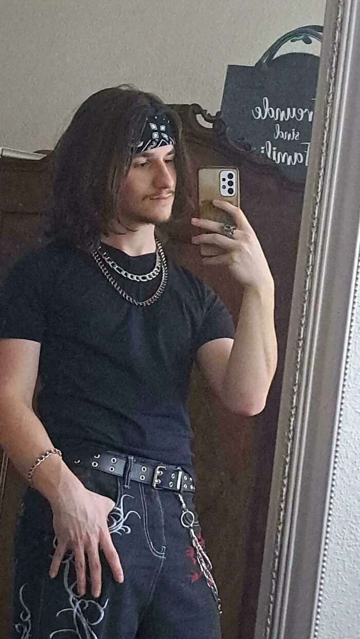 Young man with fierceflow hairstyle wearing black shirt and chains taking a mirror selfie, showcasing beautiful hair men style.