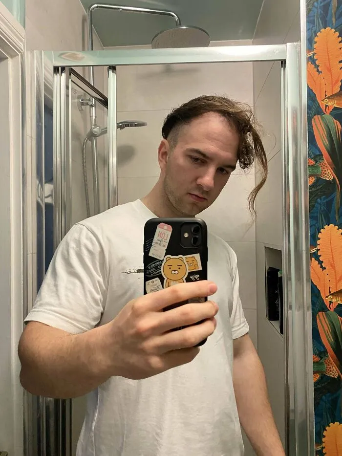 A man with an unfortunate haircut, long hair on one side and shaved on the other, looks in a mirror.