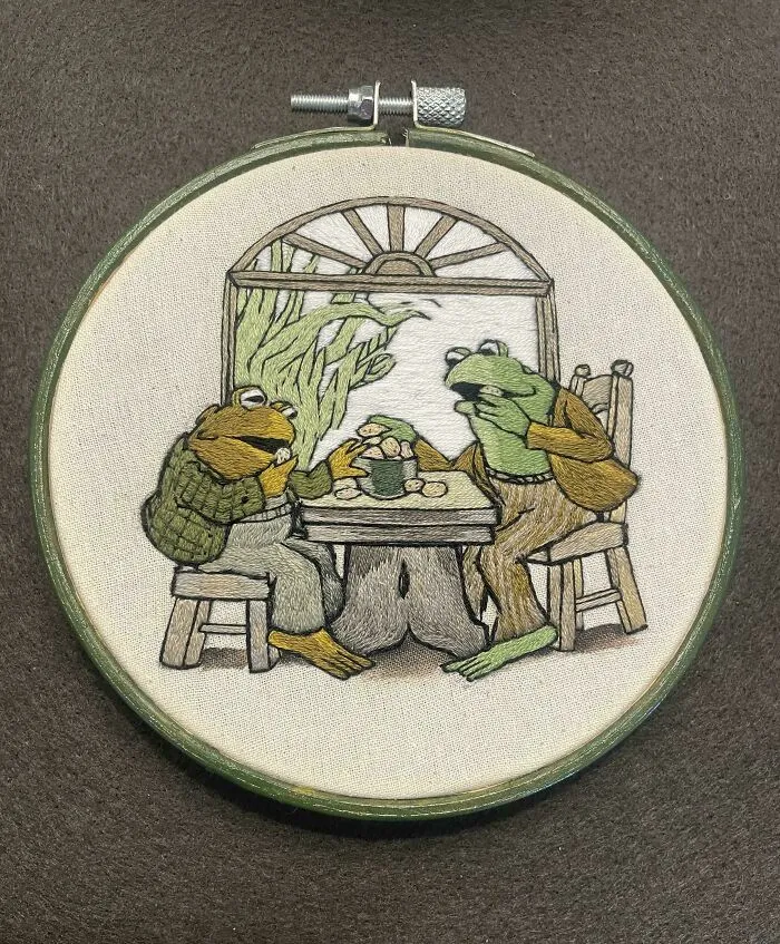 Embroidery design of two frogs sitting at a table with a window background in a wooden hoop frame.