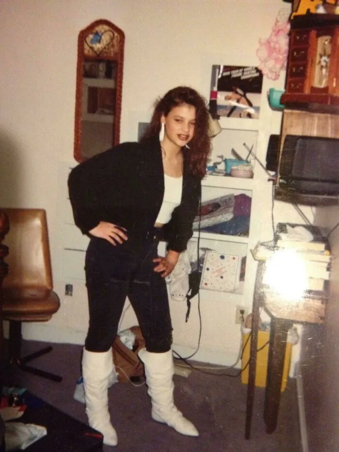 Woman dressed in 80s fashion with black jacket, leggings, and white boots, standing in a cluttered room.