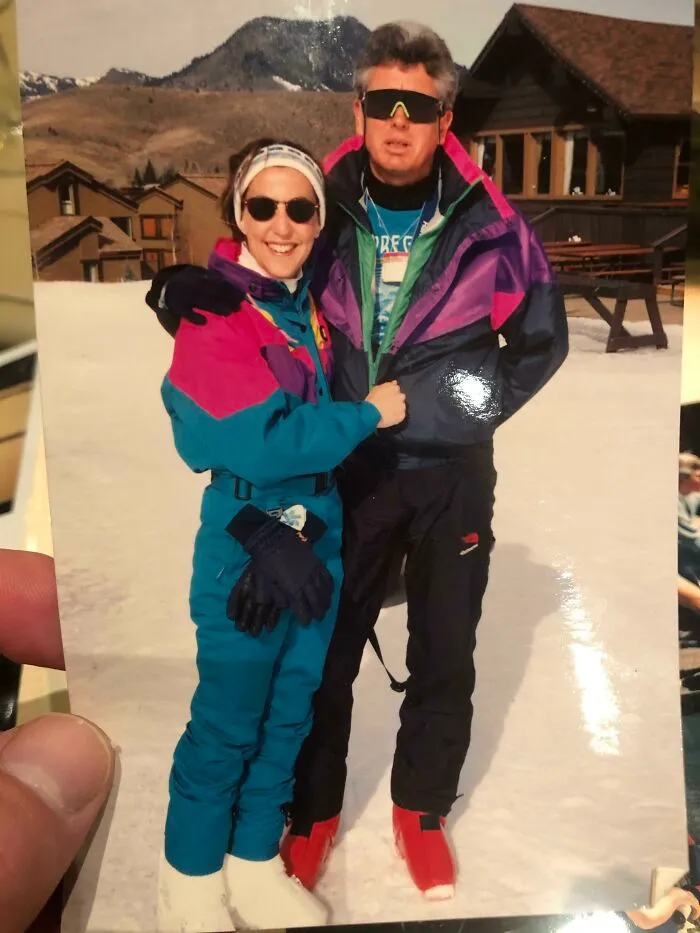Two people in colorful 80s fashion ski outfits, wearing sunglasses, posing on snow with mountains and a cabin in the background.