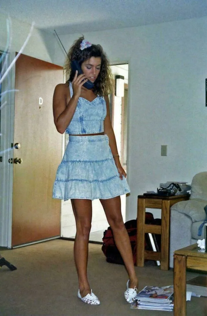 A woman in 80s fashion trends, wearing a denim outfit and talking on a vintage phone in a casual home setting.