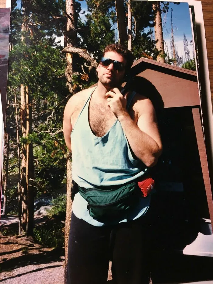 Man in 80s fashion with sunglasses, tank top, fanny pack, posing outdoors with trees in the background.