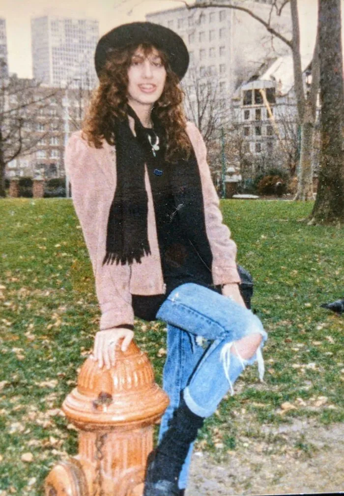 Person in 80s fashion trends, wearing a hat, scarf, and ripped jeans, posing by a fire hydrant in a park.