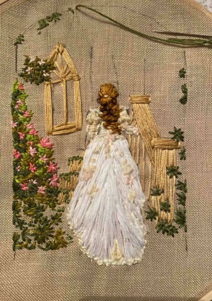 Embroidery showing a woman in a white dress with brown hair near detailed flowers and a staircase on beige fabric.