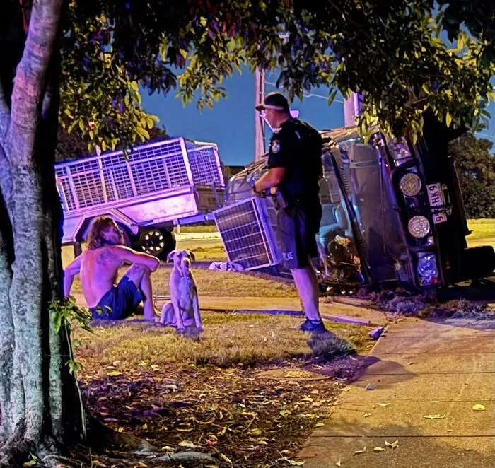 Policía interactúa con un hombre al lado de un auto volcado en Australia, con un perro presente.
