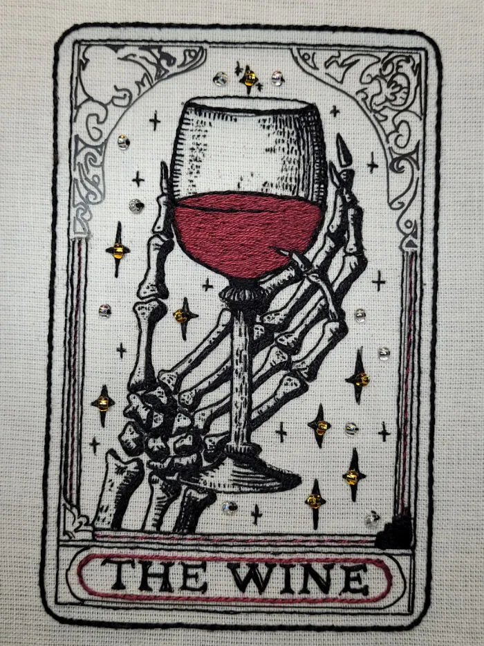Skeleton hand embroidery holding a wine glass with red wine, detailed stitching and decorative accents showcasing incredible embroidery skills.