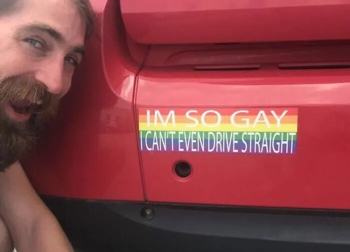 A man smiling next to a red car with a funny bumper sticker about driving.