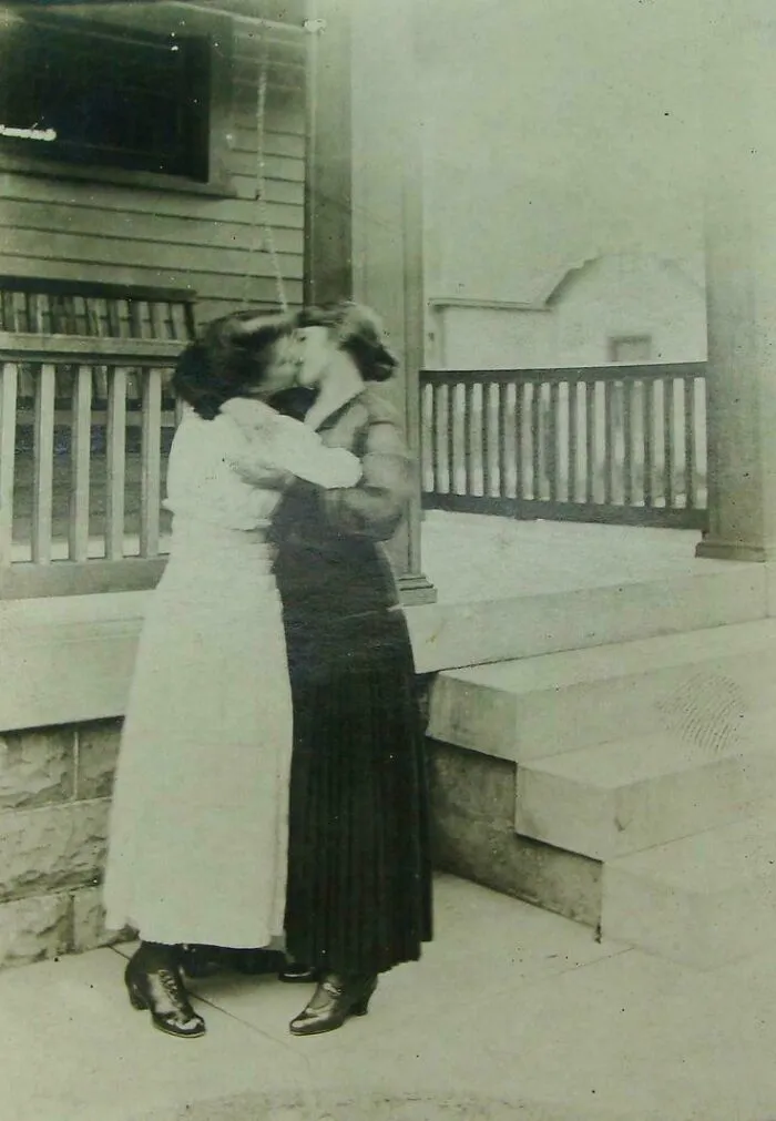 Victorian era women sharing a kiss on a porch, both wearing long skirts and boots, capturing intimacy in a historical context.