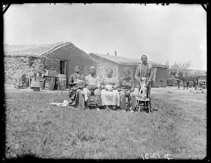 Victorian era family sitting outside a sod house, with a man standing nearby.