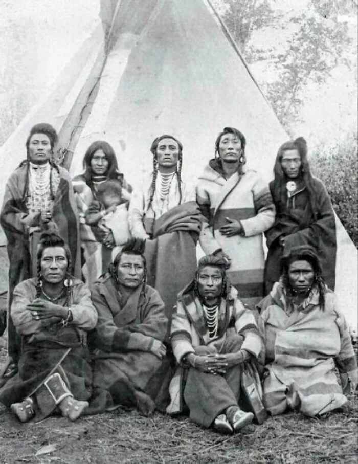Retrato grupal de hombres nativos americanos (tribu crow), una mujer y un niño frente a un tipi en Colorado, 1880