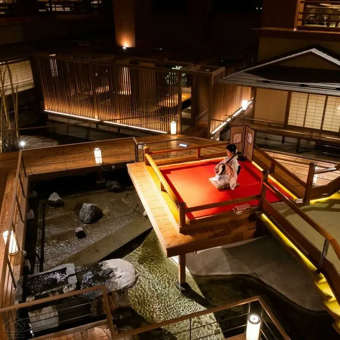 Woman in traditional attire seated on a red platform, surrounded by serene Japanese architecture at night.