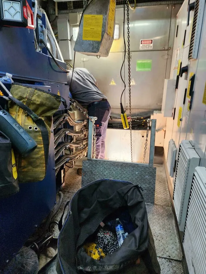 Worker ignoring safety protocol while crawling through industrial machinery in a hazardous work environment with exposed cables.