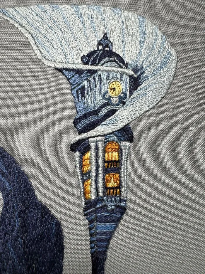 Detailed embroidery of a blue clock tower with illuminated windows showcasing beautiful embroidery skills on gray fabric background.