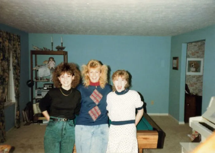 Three women in 80s fashion trends with big hair and vibrant outfits, standing in a living room.