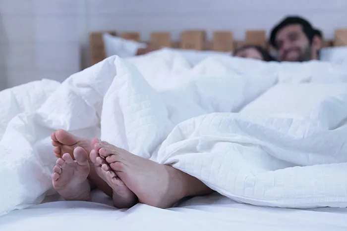Feet of a couple peeking out from under white sheets, symbolizing secrets confessed in intimate moments.