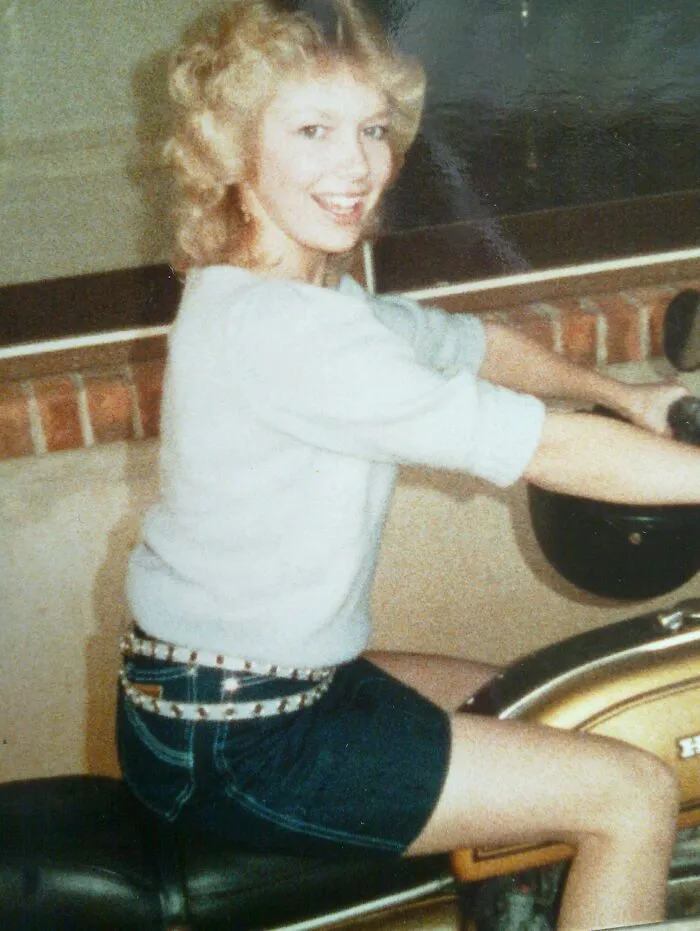 Woman in 80s fashion, wearing denim shorts and studded belt, sitting on a motorcycle indoors.