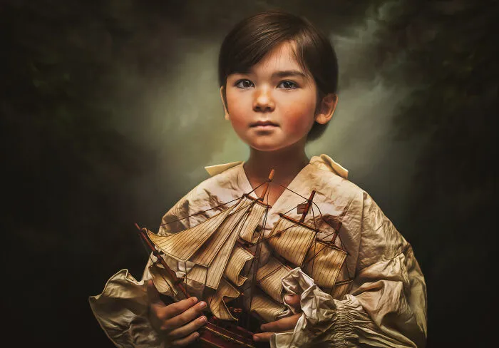 Creative photo of a child holding a model ship, wearing a vintage-style shirt against a moody background.