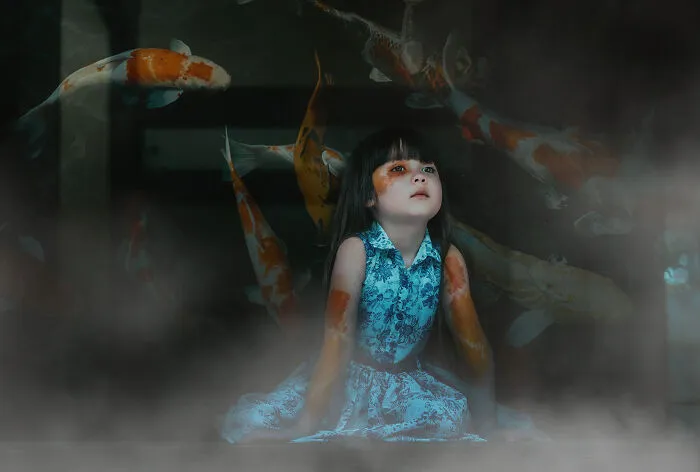 A child in a blue dress sits surrounded by koi fish, illustrating a creative photography concept.