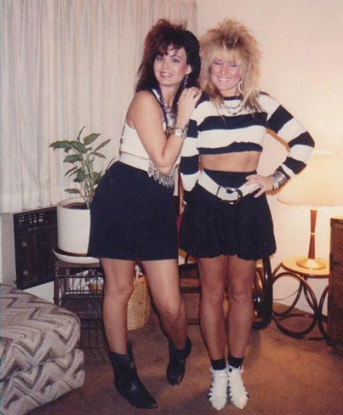 Two women in 80s fashion trends with big hair, wearing striped and black outfits, posing in a retro living room.