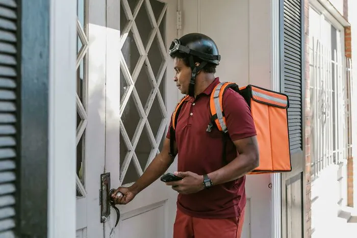 A delivery person in a helmet and uniform attempting to unlock a front door, carrying a large insulated bag.