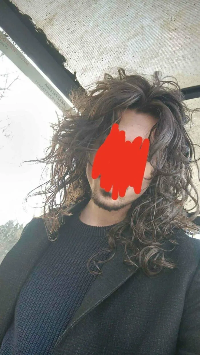Man with fierceflow long curly hair wearing a black sweater and coat in an outdoor setting with a glass roof above.