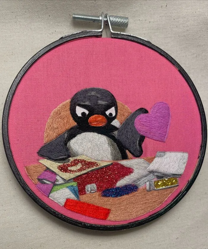 Embroidered penguin holding a purple heart with various colorful craft materials on a pink fabric background in embroidery hoop.