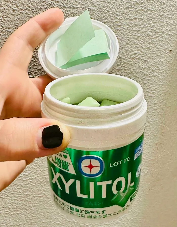A hand holding an open container of Lotte xylitol gum, revealing green square pieces.