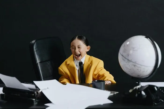 Child in a yellow jacket laughing in an office setting with scattered papers and a globe, related to jokes about office.