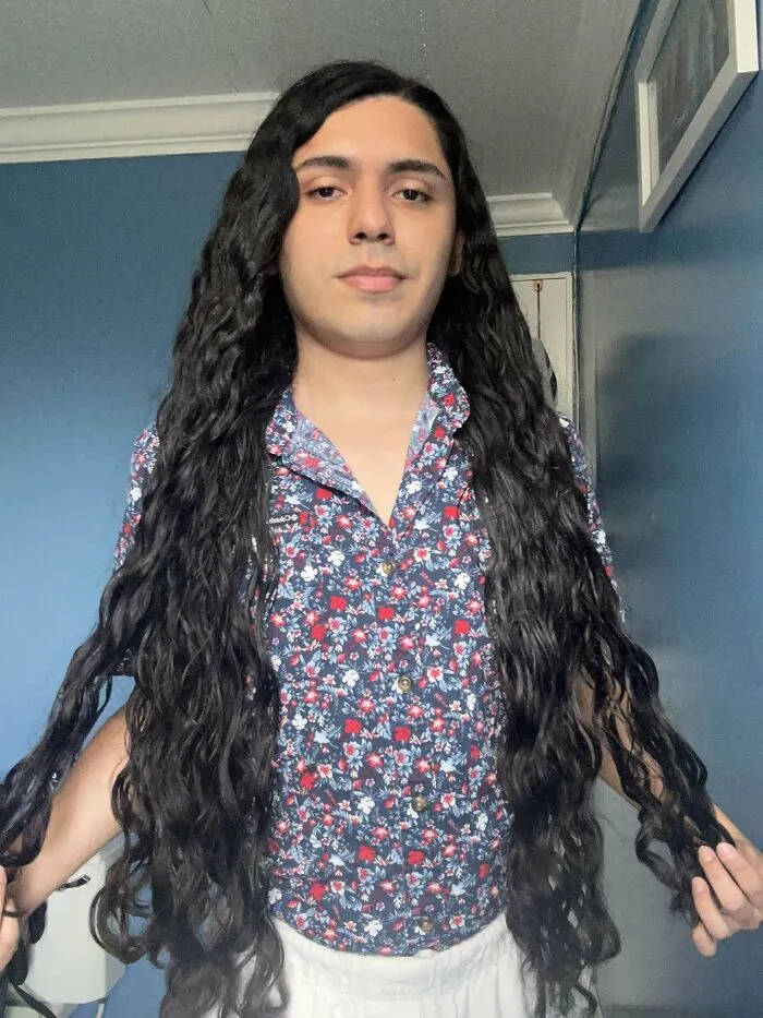 Man with fierceflow long curly hair wearing a floral shirt standing indoors against a blue wall.