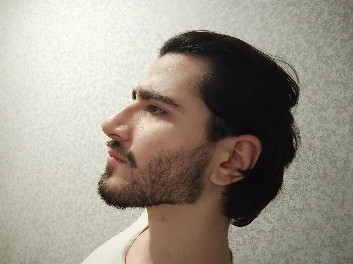 Young man with fierceflow beautiful hair, styled back, showing sharp profile against a textured light background.