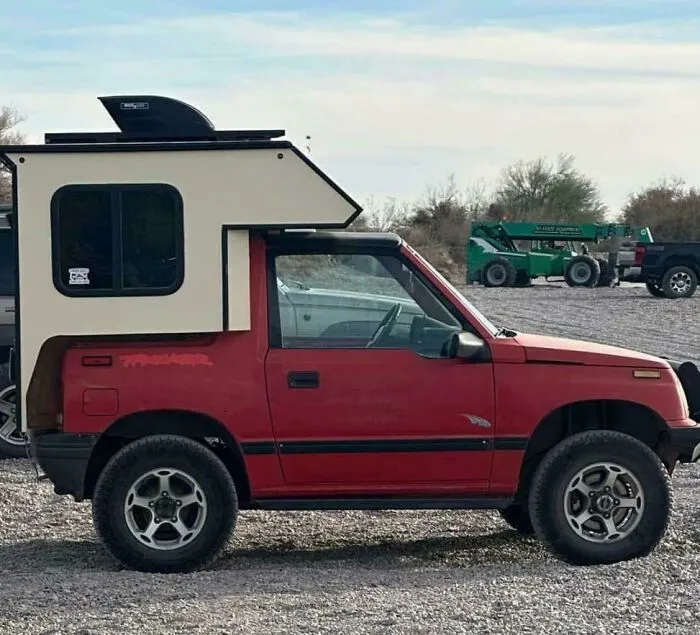 Compact red truck with a homemade camper modification parked on a gravel lot, showcasing redneck engineer creativity.