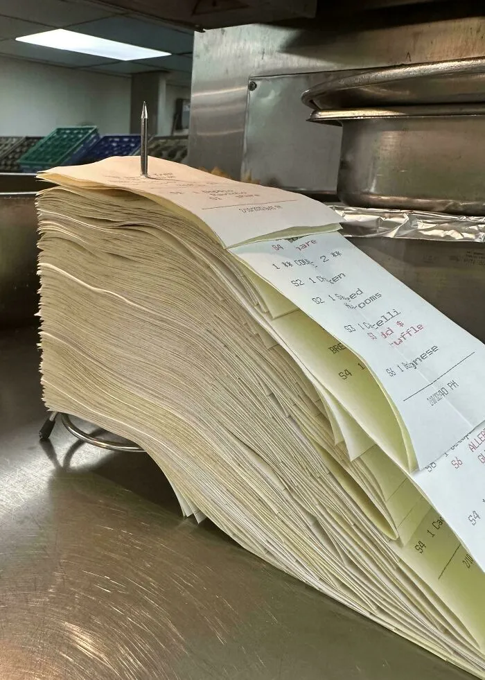 A stack of food service order tickets pierced by a spindle in a restaurant kitchen setting.