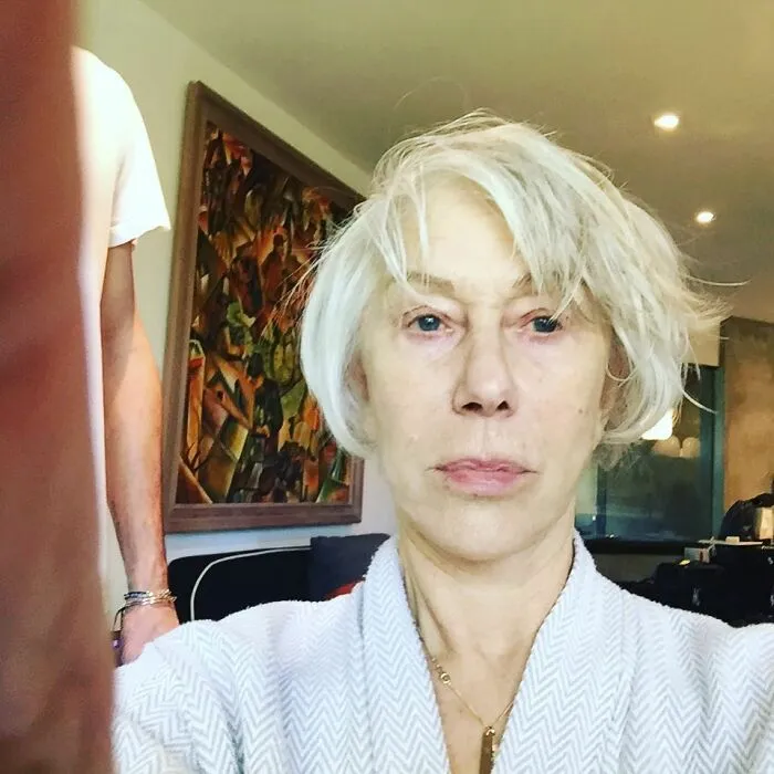 A person with short white hair wearing a robe, indoors with no makeup, standing near a painting.