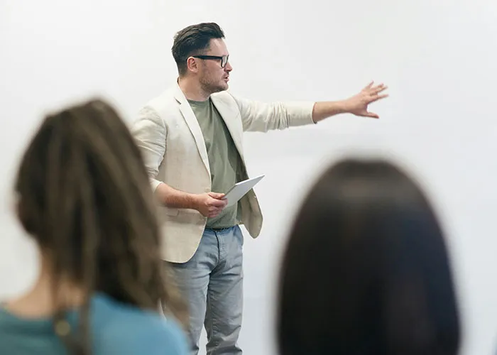 Man in a beige blazer holding a tablet, gesturing while speaking to an audience about workplace dynamics and revenge.