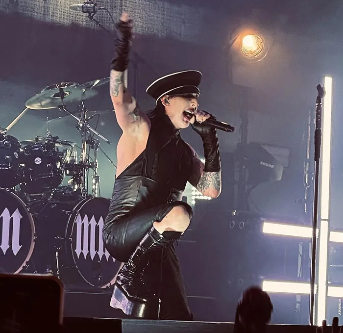 Singer performing energetically on stage in dramatic attire, with drums and bright stage lights in the background.