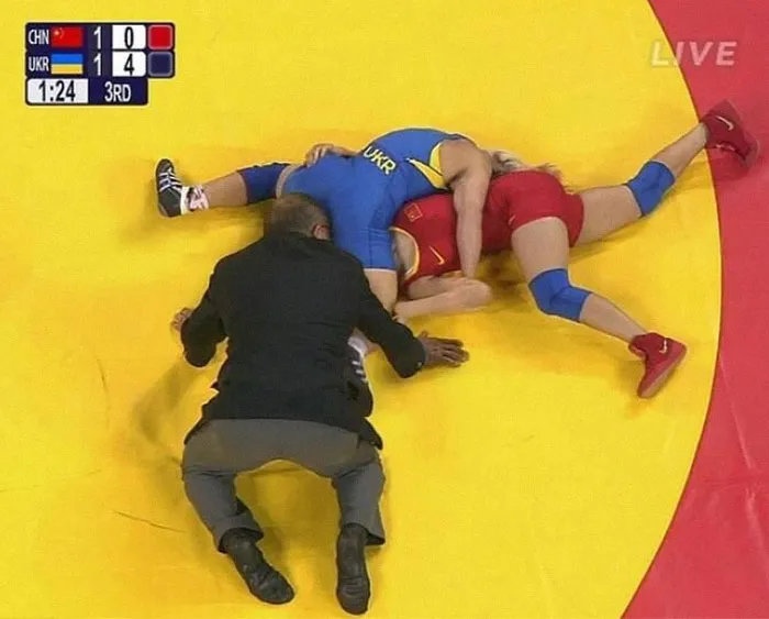 Árbitro observa un movimiento raro en un combate de lucha, atletas en el suelo, uno con uniforme azul y otro rojo.