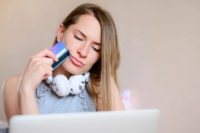 Woman pondering financial decisions, holding a credit card near a laptop with headphones around her neck.