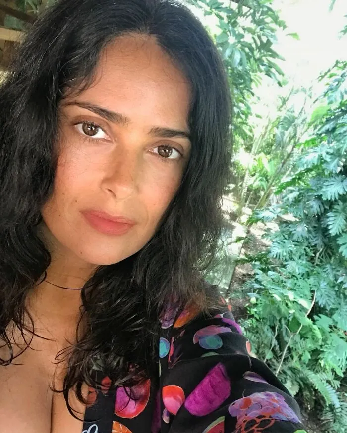 Celebrity without makeup, dark hair, floral shirt, outdoors with greenery in the background.