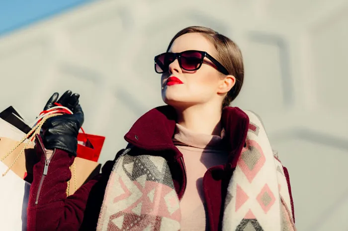 Woman in stylish coat and sunglasses holding shopping bags, illustrating the rich out of touch with reality.