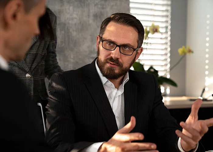 Man in a suit and glasses gesturing during a discussion in a well-lit office, emphasizing careful workplace interactions.
