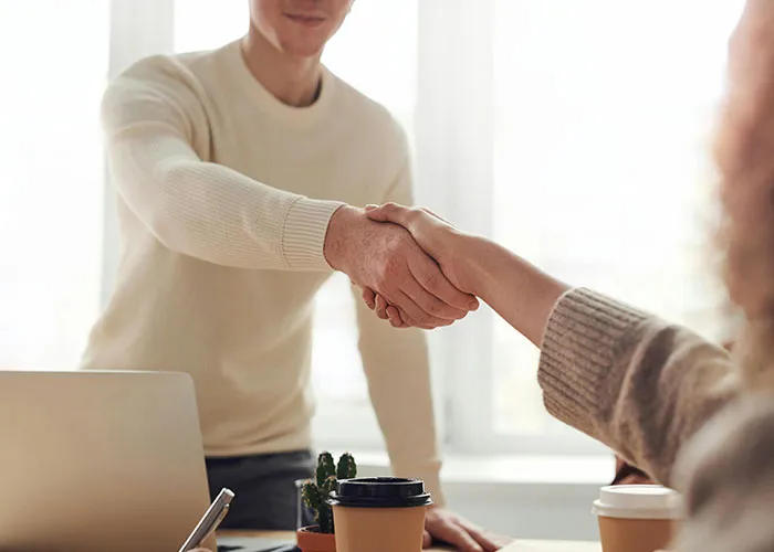 Two people shaking hands in an office setting, with a laptop and coffee cups on the table.