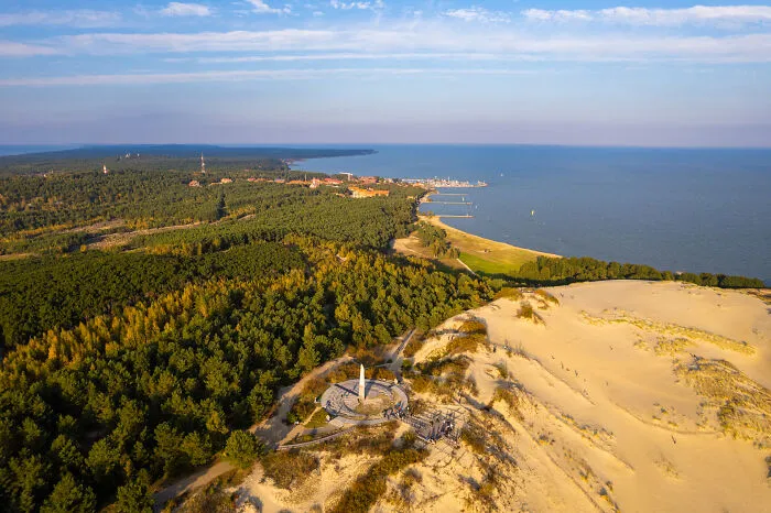 Bird’s-eye view of Lithuania showcasing sandy dunes, lush forests, and a distant coastline under a clear blue sky.