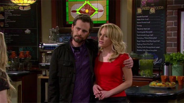 TV couple standing in a café, lacking chemistry, man in dark jacket with arm around woman in red top.